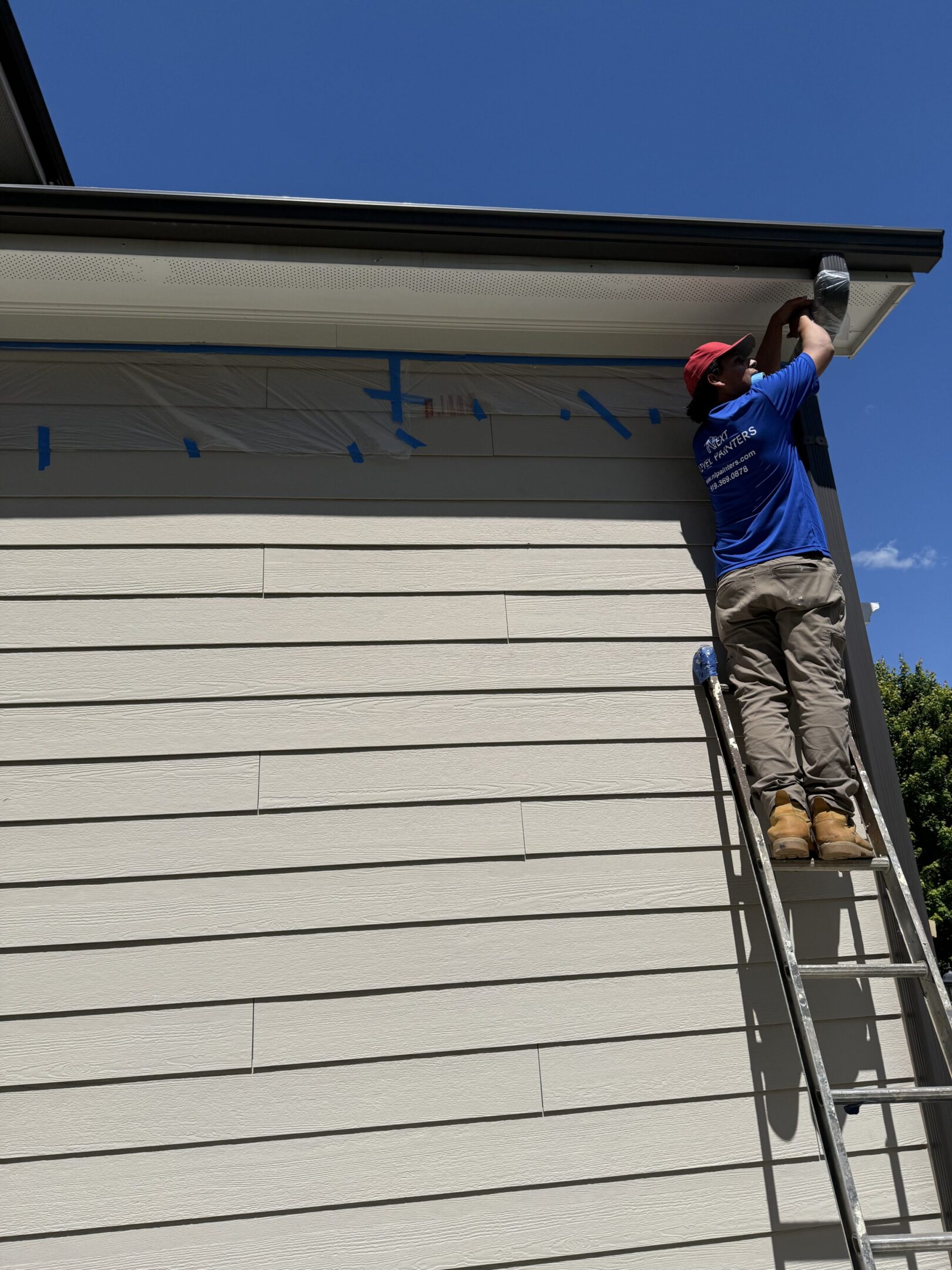 Man on ladder painting a house exterior under clear sky.
