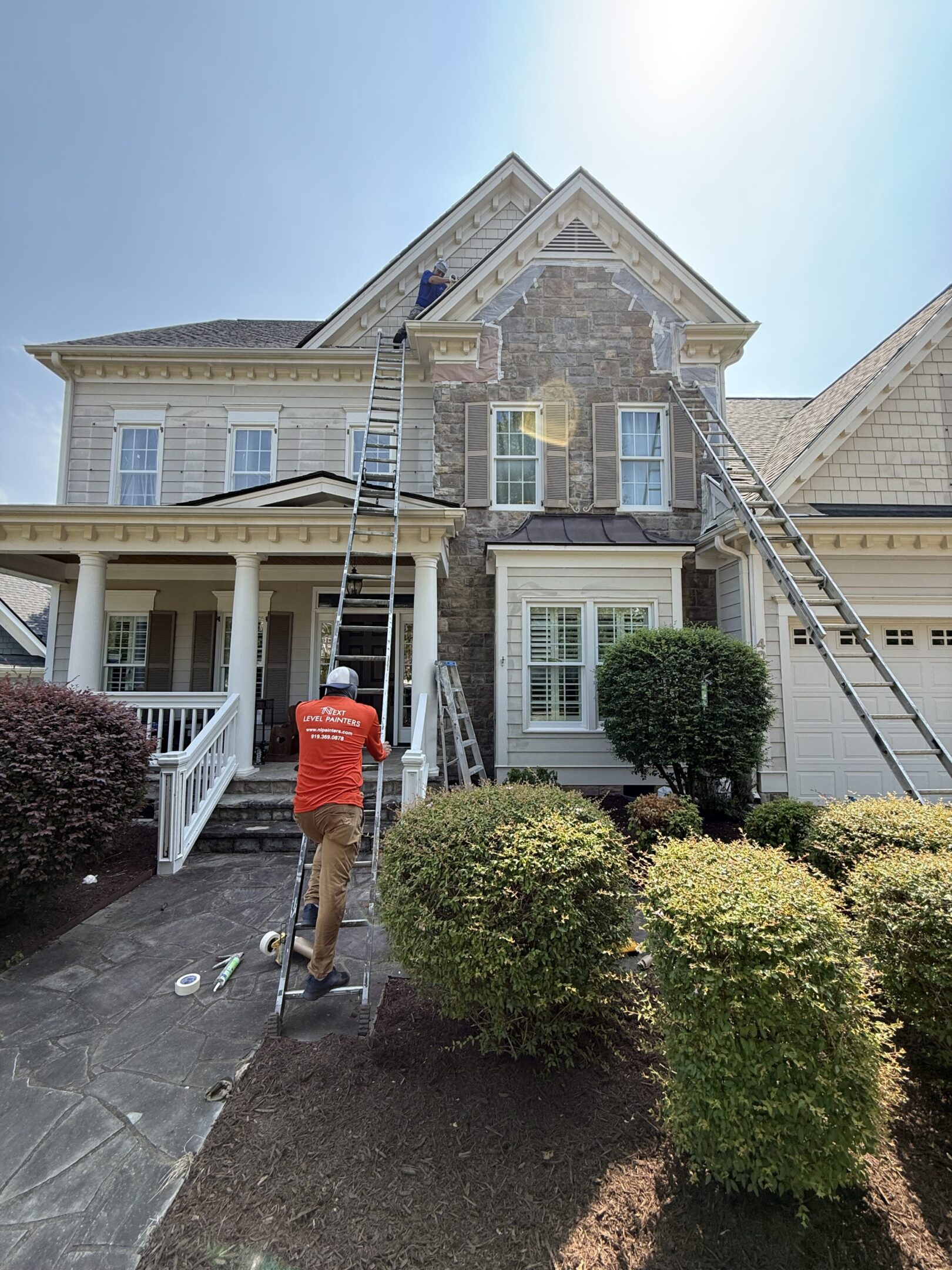 Workers painting a two-story house exterior on a sunny day.