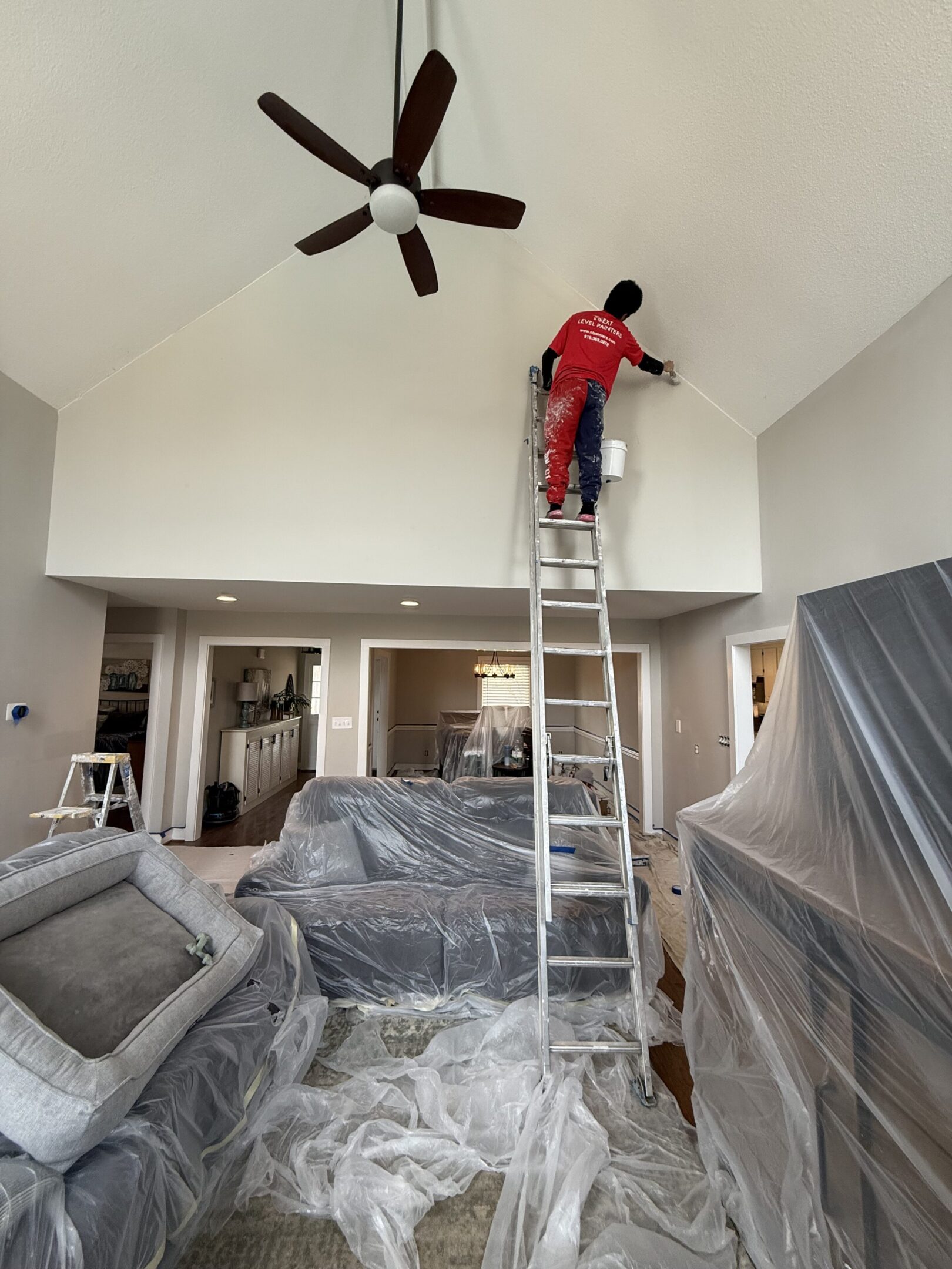 Person in red working on a high ceiling using a tall ladder in a covered living room.