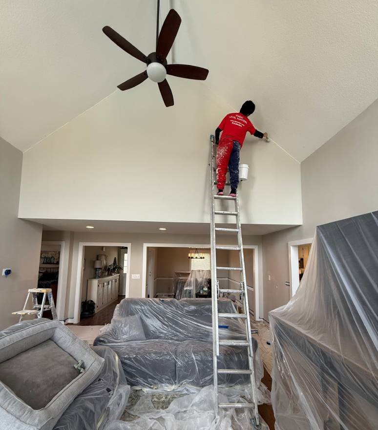A worker on a tall ladder painting a high ceiling in a living room.