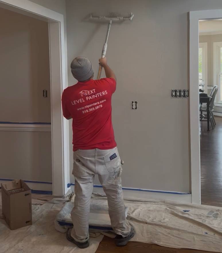 Painter applying white paint to a beige wall using a roller.