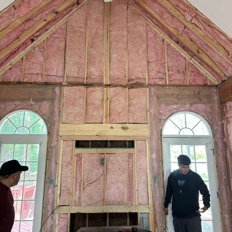 Two men stand inside a partially constructed room with exposed insulation and wall framing.